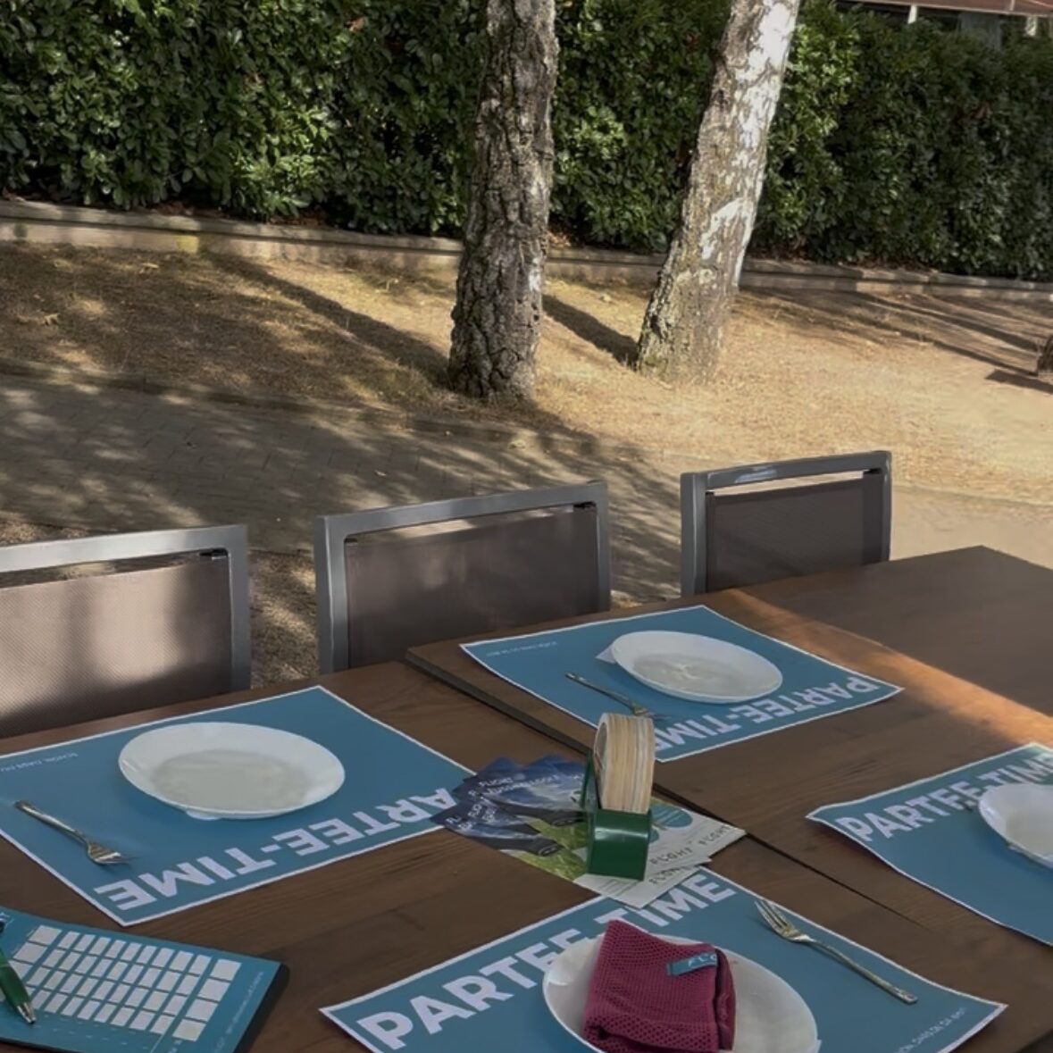 Outdoor dining table set with blue PARTY TIME placemats, white plates, and napkins under a shaded patio next to greenery and trees.